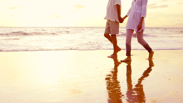 Older Couple Holds Hands and Walks Down the Beach at Sunset Getting Their Feet Wet. Slow Motion