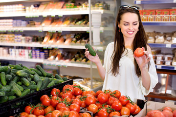 During the shopping young smiling woman is choosing cucumbers and tomatoes