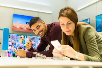 Fototapeta premium Two people are choosing smart phones. Woman is de-focused, man is in focus looking at camera holding thumb up. Shallow depth of field.