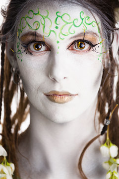Close up of a white body painted girl.
She represents an angel or an elf or a fairy tale creature.
Brown hair with dreadlocks, brown eyes and gold painted lips.