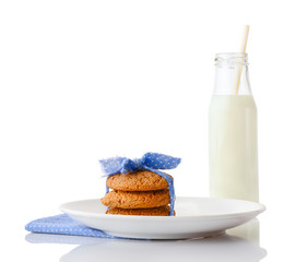Stack of three homemade oatmeal cookies tied with blue ribbon in small white polka dots on white ceramic plate on matching blue napkin and bottle of milk with straw, isolated on white background