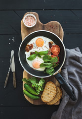 Pan of fried eggs, bacon, tomatoes with bread, mangold and cucumbers on rustic wooden serving board over dark table surface