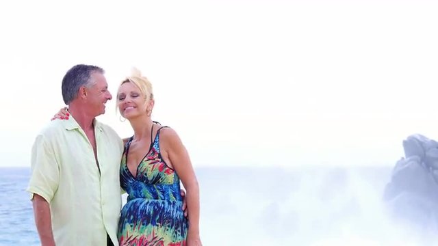An older couple kiss each other on the beach and then smile, with waves crashing on rocks behind them