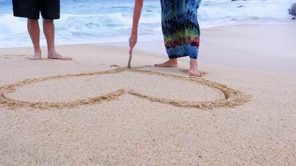 An older woman draws a heart and initials in the sand and her husband gives her a kiss - Powered by Adobe