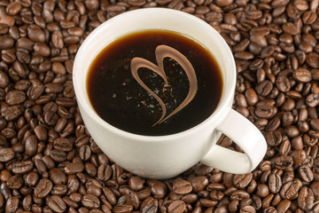 Eine weisse Kaffeetasse steht inmitten von Kaffeebohnen. Gefüllt mit Kaffee. Ein geschwungenes Herz verziert den Kaffee.