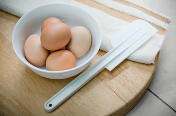 the five eggs in white ceramic bowl, white plastic spatula and brown strip white towel on wood table