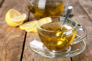 Glass cup of tea and teapot on wooden background decorated with sliced lemon and green leaves, close up