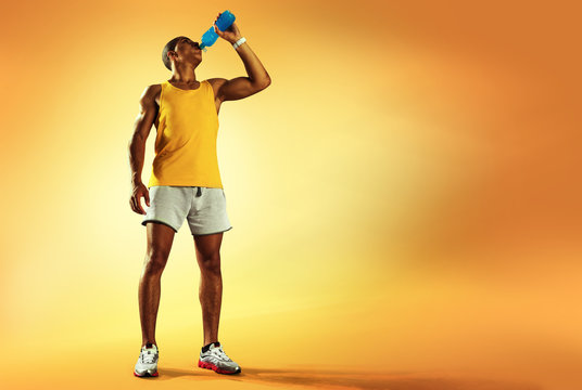 Young muscular build man drinking water of bottle after running, attractive athlete resting after workout outdoors, fitness and healthy lifestyle concept. Isolated on yellow