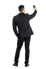 Obraz premium Rear view of young executive in black suit taking photo with his cellphone. Full body length portrait isolated over white studio background. 
