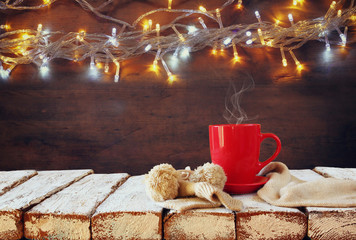Cup of hot coffee and cozy knitted scarf on wooden table in front of garland lights background
