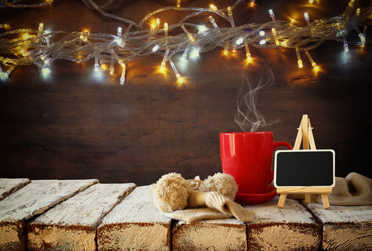 Cup of hot coffee and blackboard on wooden table in front of snowy garland lights background