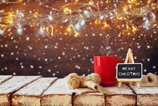 Cup of hot coffee and blackboard with words "merry christmas" written on it on wooden table in front of snowy garland lights background