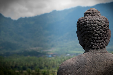 Stone Buddha statue from behind at buddhist temple Borobudur facing the Mountains