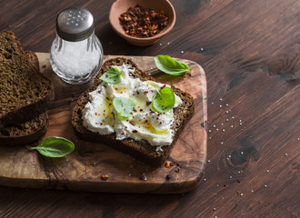 Sandwich with soft cheese, olive oil and basil, served on olive cutting board on dark wooden surface. Healthy Breakfast or snack