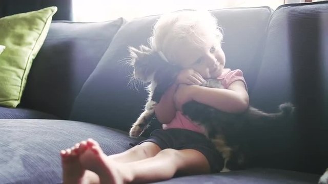 A little girl sitting on the couch holding a kitten and giving it a hug