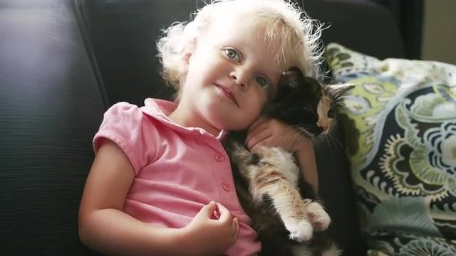 A little girl sitting on the couch holding a kitten and giving it a hug