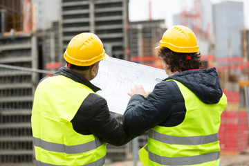 Back turned construction workers with yellow hardhat and safety jacket checking blueprint