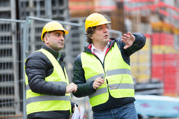 Architect and construction worker with yellow safety jacket and hardhat talking at construction site