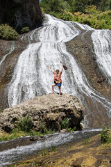 Young girl with hands up, with ukulele, standing on a rock near waterfall