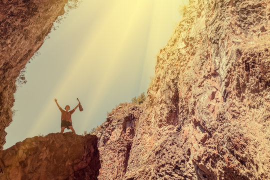 Silhouette of a young girl with hands up, with ukulele, standing on a rock near the cave against the sky