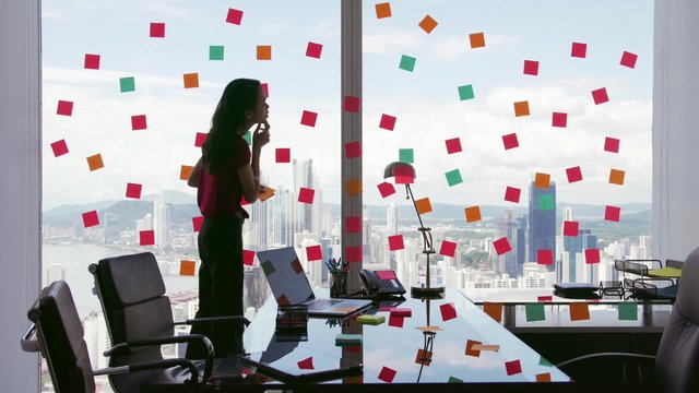 Latin american woman working as secretary in modern office with beautiful sight on the city, sticking adhesive notes with tasks on skyscraper window. The girl feels overwhelmed. Wide shot