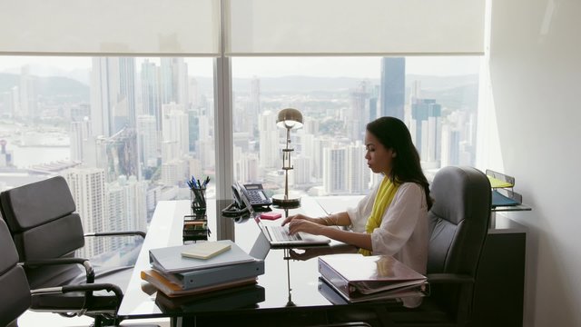 Adult businesswoman sitting in modern office with beautiful sight of the city. The secretary writes fast on laptop pc with serious expression. Wide shot