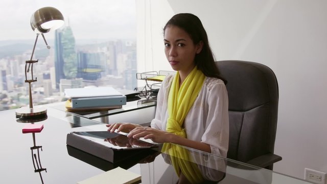Adult businesswoman sitting in modern office with beautiful sight of the city. The secretary writes on tablet pc, then turns to camera and smiles. Slow motion, steadicam shot