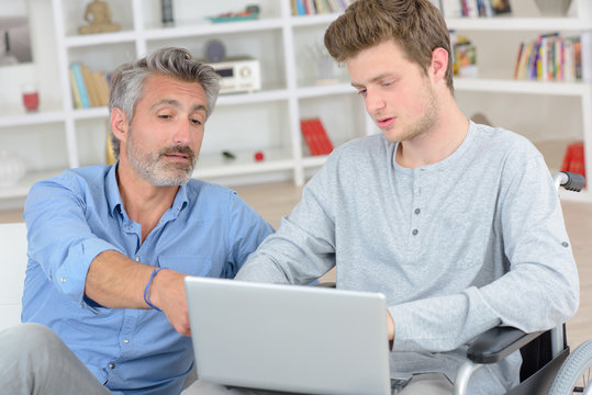 Father pointing to screen of laptop held by son in wheelchair