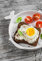 Sandwich with feta cheese and boiled egg, tomatoes, and basil on a white plate on a wooden surface. Healthy breakfast or snack