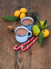 Hot chocolate in enamel metal mugs, fresh mandarins, cinnamon sticks, pine cone and candy canes over rustic wooden background