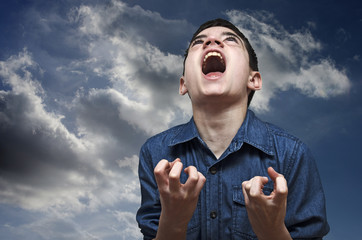 Boy screaming.Angry caucasian teenager screaming wildly on a cloudy sky background.