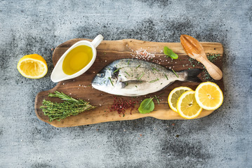 Fresh uncooked dorado or sea bream fish with lemon, herbs, oil and spices on rustic wooden board over grunge backdrop