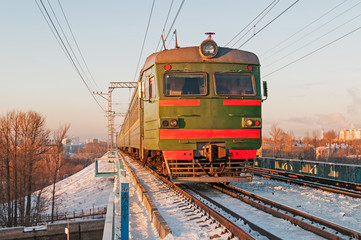 Green suburban electric train moves towards on snow-bound railroad vanishing in horizon before sunset light against skyline background. Moscow, Russia. 
