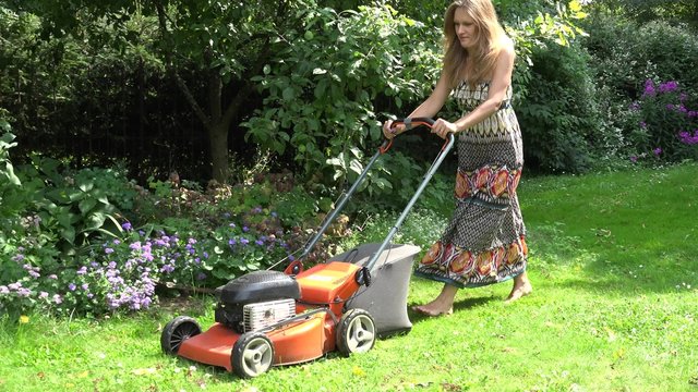 pretty blond woman start grass lawn cutter and push near flower bed in backyard. Girl spend leisure landscaping garden environment in summer time. Panorama shot. 4K