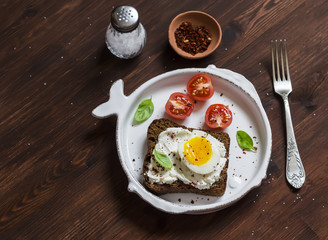Sandwich with feta cheese and boiled egg, tomatoes, and basil on a white plate on a dark wooden surface. Healthy breakfast or snack
