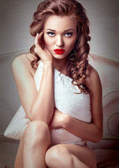 Beauty Sexy Woman Model Girl Sitting on Bed, Touching her Face with Hand and Covering with Pillow. Long Curly Hair, Red Matte Lips, Blue Eyes