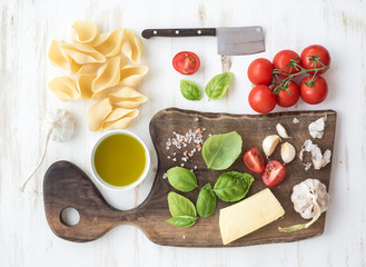 Ingredients for cooking pasta. Conchiglioni, basil leaves, cherry-tomatoes, Parmesan cheese, olive oil, salt, garlic on rustic walnut chopping board over white wooden background