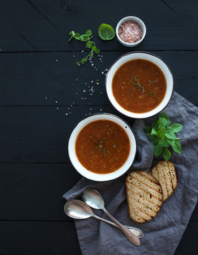 Roasted tomato soup with fresh basil, spices and bread in rustic metal bowls over black background