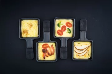 Fotobehang Four small square raclette pans with melted cheese and different ingredients: paprika, bacon, cherry tomatoes, broccoli and pear slices © inats