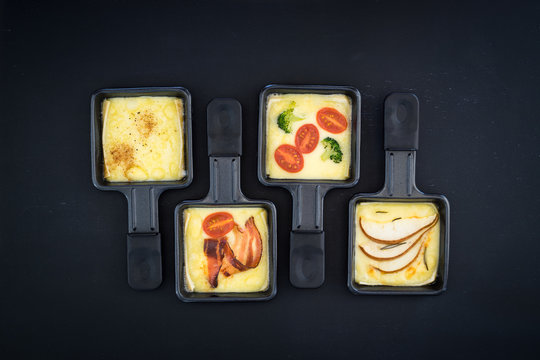 Four small square raclette pans with melted cheese and different ingredients: paprika, bacon, cherry tomatoes, broccoli and pear slices