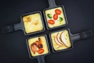Gordijnen Four small square raclette pans with melted cheese and different ingredients: paprika, bacon, cherry tomatoes, broccoli and pear slices © inats