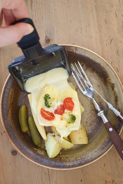 Pouring melted raclette cheese from square raclette pan onto boiled potatoes and pickles