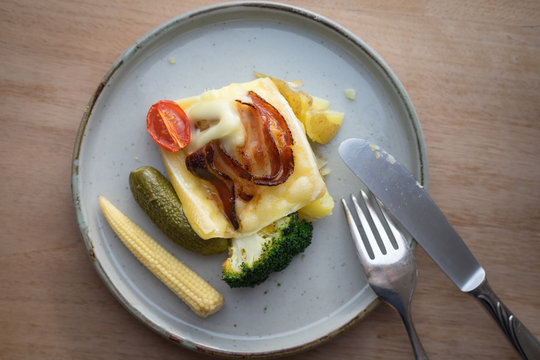 Plate with boiled potatoes, broccoli and pickles covered with melted raclette cheese with grilled bacon slice