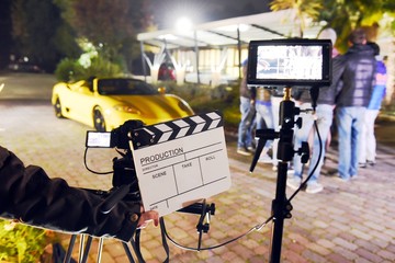 Operator holding clapperboard during the production of short film outdoor in the night with sportive yellow car and actor on stage. Focus on the clapperboard and monitors - obrazy, fototapety, plakaty