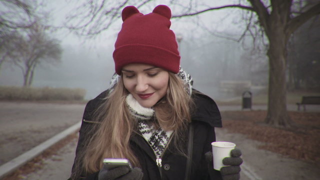 Woman in red hat looking at phone and smiling. Young blonde woman holding coffee in autumnal park.