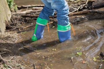 Ein Kind in blauen Gummistiefeln läuft durch einen Flusslauf. Im Hintergrund ein Staudamm aus Baumästen. - 97707010