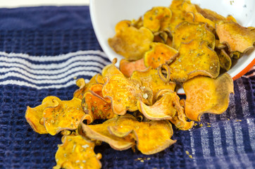
    Bowl of homemade baked sweet potato chips poured on a blue kitchen towel and raw vegetable on a cutting board.Healthy sweetpotato vegetarian crisp snack seasoned with oregano,olive oil.