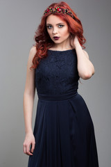 Beautiful red-haired fashion model posing in evening dress and in the diadem over dark background. female gestures of seduction. body language. touching her hair