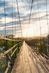 Obraz premium Old vanishing lengthy hanging wooden footbridge with rails over river and bright Sun in sky at sunset. Arkhangelsky region, Russia. 