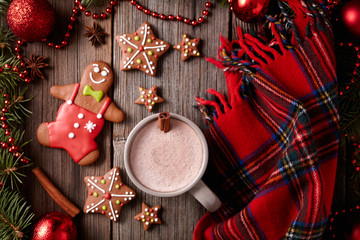 Cup of hot chocolate with gingerbread man, stars cookies and warm scarf composition in fir tree decorations frame on vintage wooden table background. Homemade traditional dessert recipe.
